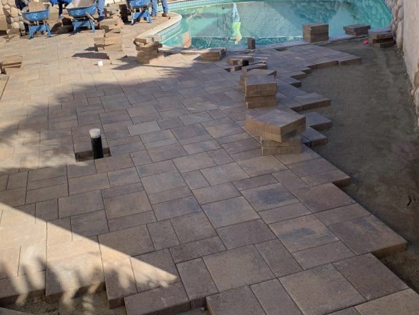 Crew laying a paver pool deck with stacks of pavers ready to go in