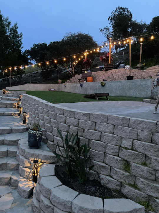 Finished terraced backyard with stone retaining walls, paver patio, turf and string lights at dusk